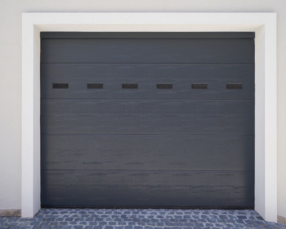 A Modern Dark Grey Sectional Garage Door With Small Rectangular Windows, Highlighting Contemporary Types Of Garage Doors Suited For Energy Efficiency. This Project Was Masterfully Led By Contractor Dmytro Shevchenko From Prime Garage Door Repair. The Door Is Framed By A Crisp White Trim, Demonstrating A Stylish Contrast That Works Well For Renovated Homes In The Chicago Area.