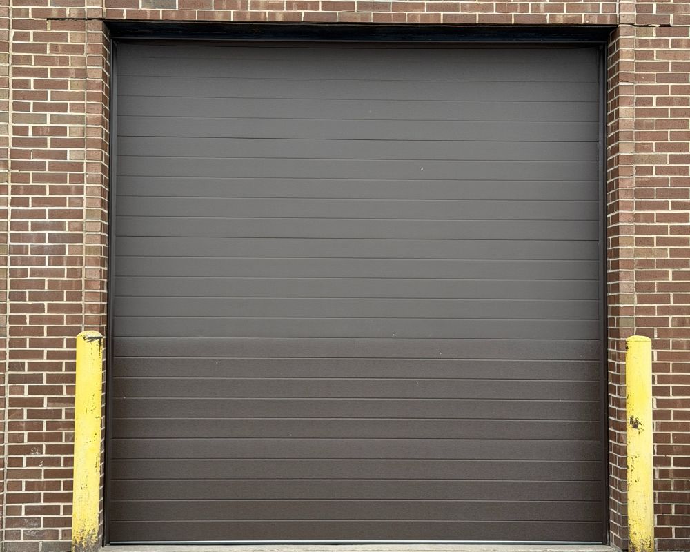 A Professional Installation Of A Dark Brown Ribbed Sectional Door, Illustrating Different Types Of Garage Doors For Commercial Or Residential Use. The Project Was Led By Contractor Dmytro Shevchenko From Prime Garage Door Repair. The Door Is Set Within A Classic Red Brick Facade, Flanked By Yellow Safety Bollards, Highlighting A Clean And Durable Finish.