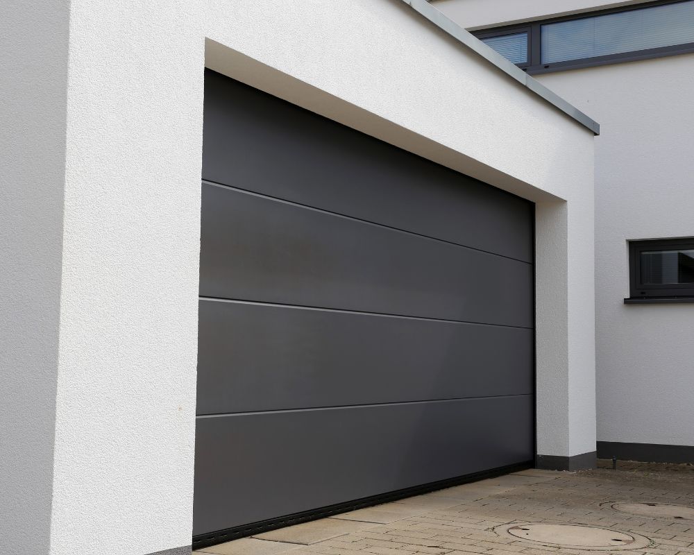 A Sleek, Wide Charcoal Grey Sectional Door On A Modern Minimalist Home, Representing High-End Types Of Garage Doors Available For Local Homeowners. The Project Was Led By Contractor Dmytro Shevchenko Of Prime Garage Door Repair. The Smooth Panels And Clean Lines Provide A Sophisticated Look, While The Durable Construction Ensures Long-Term Performance Against Heavy Snow And Wind.