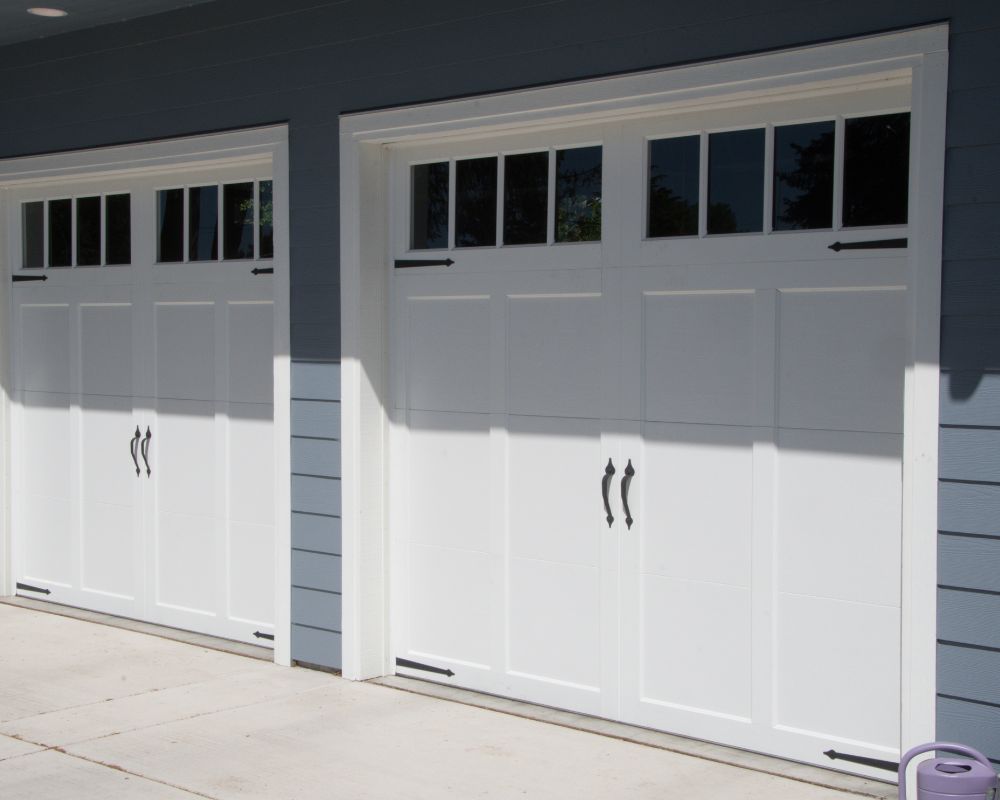 A Pair Of Elegant White Carriage-Style Garage Doors With Window Inserts, Representing Popular Types Of Garage Doors For Modern Home Aesthetics. The Project Was Masterfully Led By Contractor Dmytro Shevchenko From Prime Garage Door Repair. The Doors Feature Decorative Black Hardware And Are Integrated Into A Stylish Blue-Grey Siding Exterior, Enhancing The Home'S Curb Appeal.