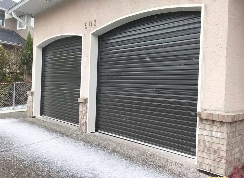 Two Elegant Black Arched Garage Doors With A Horizontal Ribbed Pattern On A Light-Colored Home During A Light Snowfall, Showcasing The Modern Garage Door Options Provided By Lead Contractor Dmytro Shevchenko In The North West Chicago Area.
