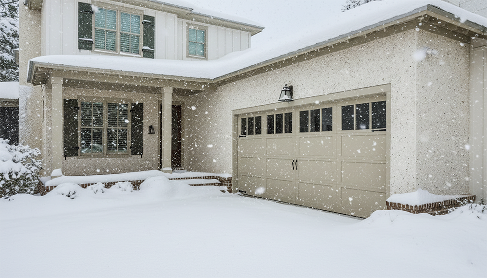 A Picturesque Home In North West Chicago Blanketed In Fresh Snow, Featuring A Tan-Coloured Panelled Garage Door With Windows Along The Top, Showcasing Attractive Garage Door Options. The House Has Textured Light-Coloured Stucco And Dark Shutters On The Windows, And The Project Was Expertly Completed By Lead Contractor Dmytro Shevchenko. Snow Covers The Ground, Porch, And Roof, With Visible Falling Snowflakes.