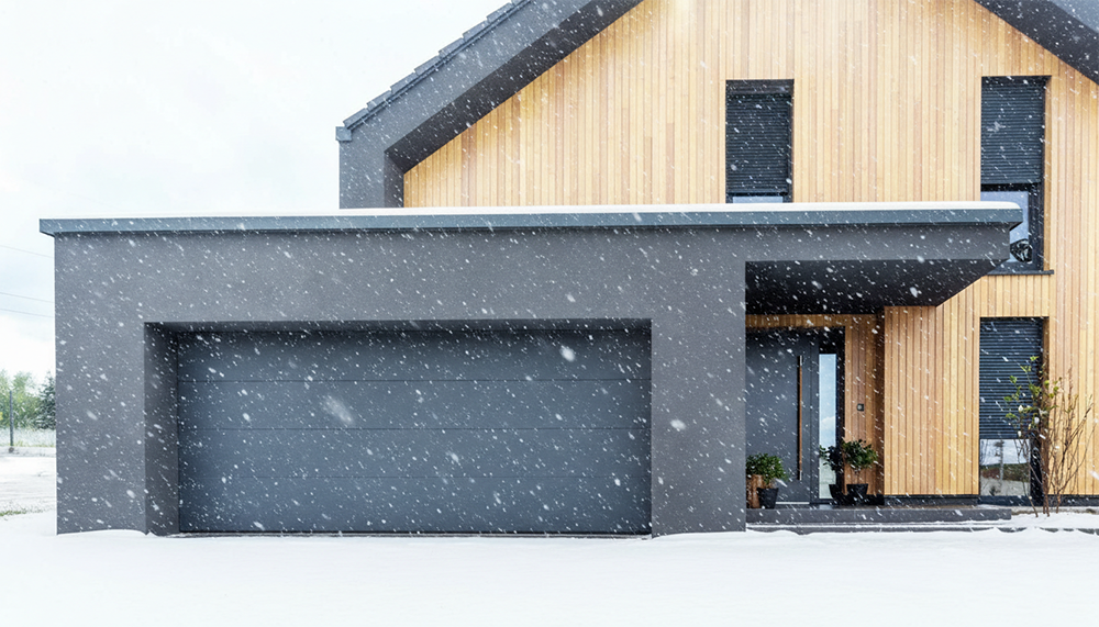 A Sleek, Modern Grey Residential Garage Door On A Contemporary Home With Wood Paneling During A Snowfall, Showcasing Premium Garage Door Options Provided By Lead Contractor Dmytro Shevchenko In The North West Chicago Area.