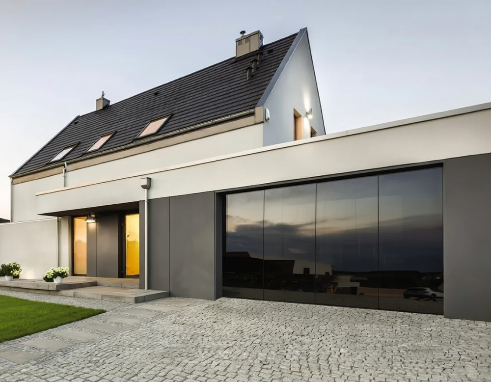 Modern Single-Family House With Dark Gray Garage Door, Large Glass Panels, And Sleek Minimalist Facade At Dusk, North West Chicago. Lead Contractor Dmytro Shevchenko Project Featuring Contemporary Architecture With Black Tiled Roof And Illuminated Entrance.