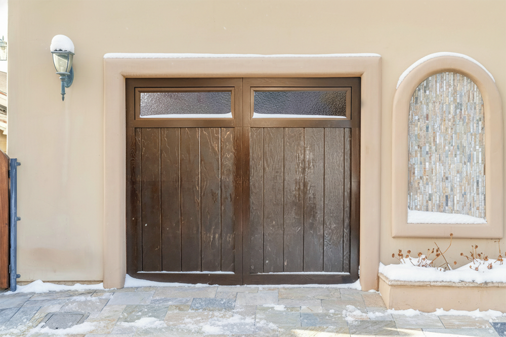 A Custom Dark Wood-Finish Garage Door With Vertical Planks And Textured Upper Windows, Installed On A Beige Stucco Home Amidst A Winter Setting. This Design Highlights The Stylish And Durable Garage Door Options Available From Lead Contractor Dmytro Shevchenko For Homeowners In North West Chicago.