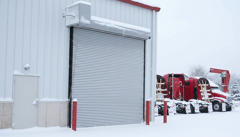 A Durable Silver Commercial Roll-Up Garage Door On An Industrial Warehouse In The Snow, Showcasing The Heavy-Duty Garage Door Options Provided By Lead Contractor Dmytro Shevchenko For Businesses In North West Chicago.