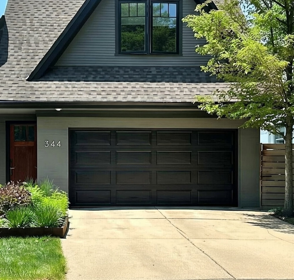 Sleek Dark Charcoal Black Double Garage Door With Raised Rectangular Panels And Modern Wood-Grain Texture, Installed On A Contemporary Gray Siding Home In North West Chicago. Garage Door Installation By Lead Contractor Dmytro Shevchenko.