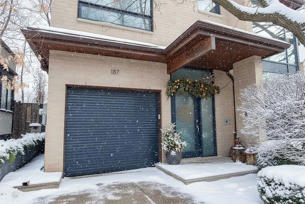 A Contemporary Home In North West Chicago During Winter, Featuring A Stylish Grey Roller Garage Door, Presenting One Of Many Sleek Garage Door Options. The Front Entrance Is Adorned With Festive Holiday Garlands. The House Has Light Brickwork, Large Dark-Framed Windows, And Is Covered With A Fresh Layer Of Snow. This Project Highlights The Work Of Lead Contractor Dmytro Shevchenko.