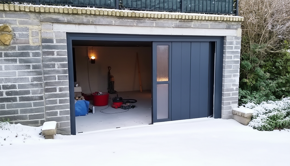 A Modern Dark Grey Side-Sliding Garage Door With Vertical Window Panels, Partially Open To Reveal The Garage Interior. The Garage Is Built Of Grey Brick And Is Surrounded By Snow, Showcasing Durable Garage Door Options For Winter Climates Provided By Lead Contractor Dmytro Shevchenko In North West Chicago.