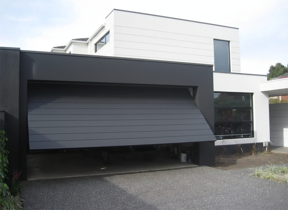 A Sleek, Modern Charcoal Grey Tilt-Up Garage Door With Horizontal Panels Partially Open On A Contemporary Minimalist Home, Showcasing High-End Garage Door Options Provided By Lead Contractor Dmytro Shevchenko In The North West Chicago Area.
