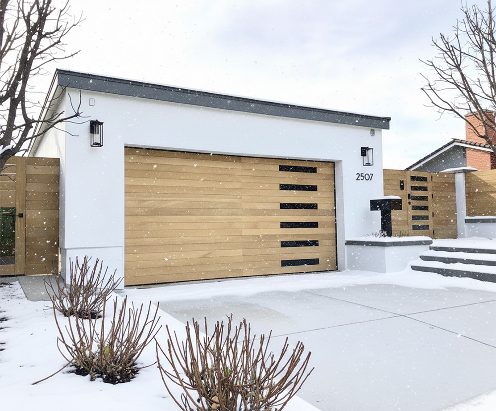 A Modern White Garage Featuring A Stylish Light Wood-Tone Garage Door With Horizontal Black Window Accents, Captured During A Light Snowfall. This Contemporary Design Highlights The Custom Garage Door Options Available Through Lead Contractor Dmytro Shevchenko For Homes In North West Chicago.