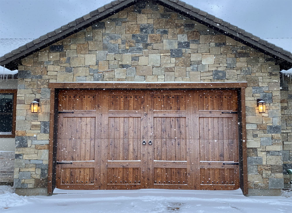 A Beautiful Custom Wood Carriage-Style Garage Door With Decorative Iron Hardware, Set Against A Stone Facade While Snow Falls. This Rustic Winter Scene Demonstrates The Premium Garage Door Options Available Through Lead Contractor Dmytro Shevchenko For Homes In North West Chicago.