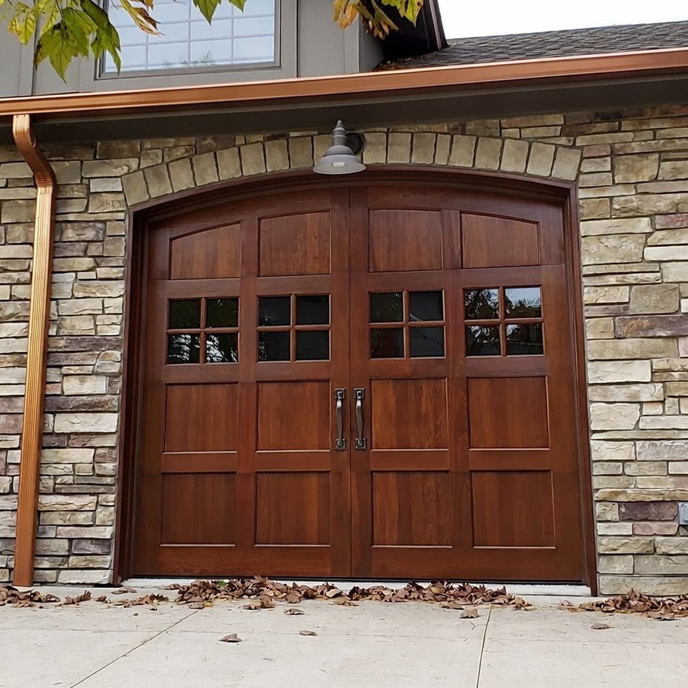 Arched Carriage-Style Double Garage Door In Rich Mahogany Wood Finish With Raised Panel Design And Upper Grid Glass Windows, Black Hardware Accents, Installed On A Stone-Front Home With Copper Gutters In North West Chicago. Garage Door Installation By Lead Contractor Dmytro Shevchenko.