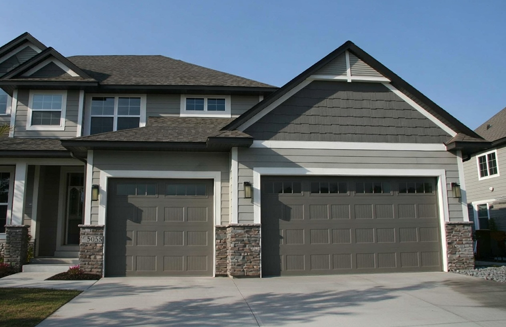 Double Garage Door In Modern Taupe Gray With Raised Panel Design And Small Rectangular Glass Inserts, Installed On A Two-Story Gray Siding Home With Stone Accents In North West Chicago. Garage Door Installation By Lead Contractor Dmytro Shevchenko.