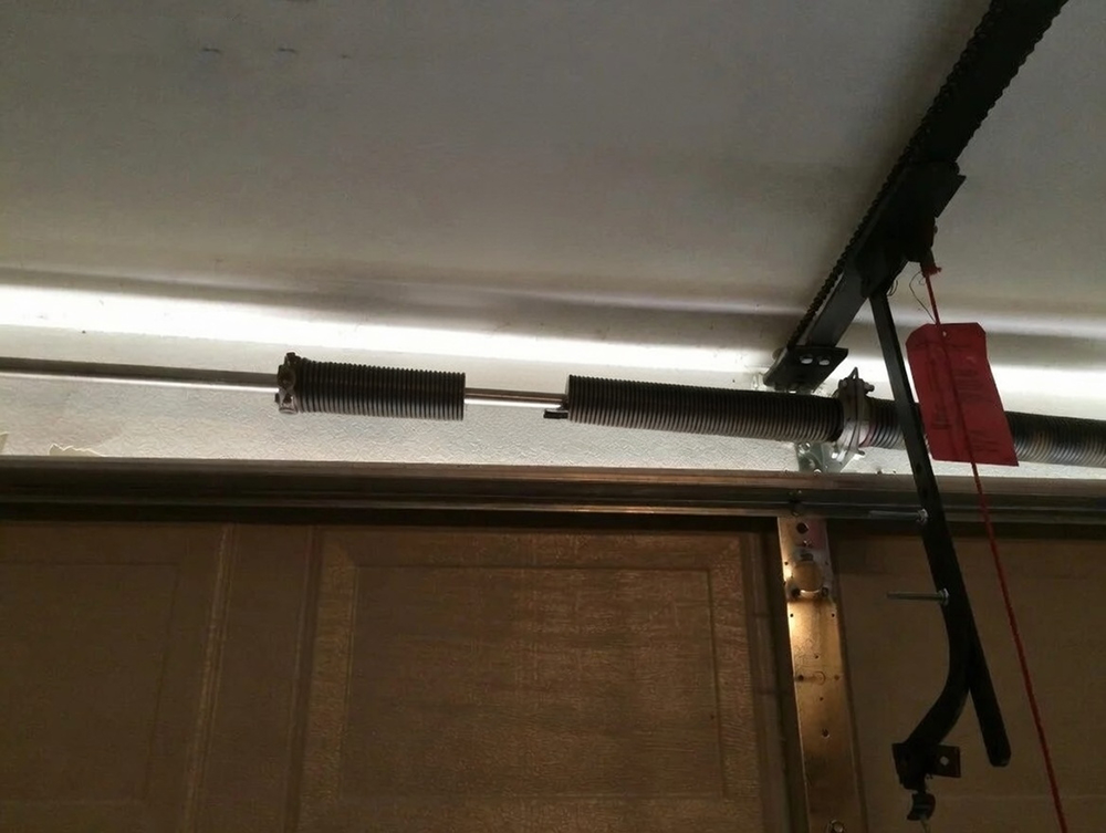 What Is A Garage Door Called? A Guide To Names And Terminology Interior View Of A Garage Door Broken Extension Spring System Mounted Horizontally Above The Door Opening, Showing The Coiled Extension Spring, Tension Cable, Pulley, And Rail Assembly In A Residential Overhead Garage Door Setup, Installed By Lead Contractor Dmytro Shevchenko In North West Chicago.