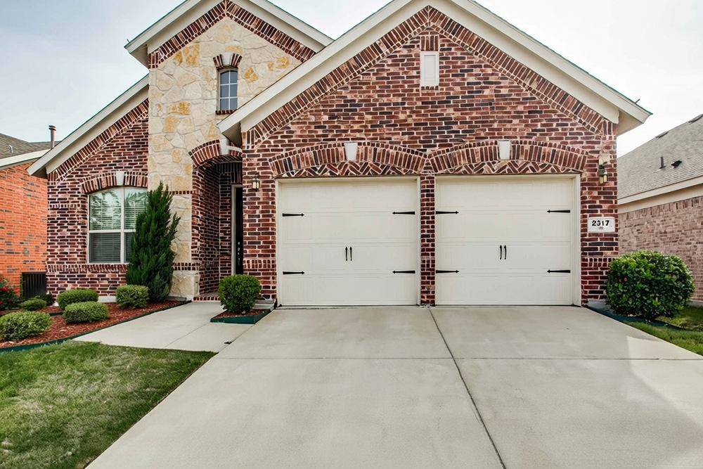 Traditional Garage Door Styles: The Timeless Choice For 2026 Classic Traditional Garage Door Styles Featuring Double White Carriage-House Swing-Out Doors With Vertical Board-And-Batten Panels, Black Wrought-Iron Strap Hinges And Handles, Arched Brick Detailing Above On A Luxurious Two-Story Red Brick And Stone Home With Gabled Roof, Address 2317 Visible, Landscaped Front Yard And Concrete Driveway, North West Chicago. Lead Contractor Dmytro Shevchenko – Timeless Traditional Garage Door Styles.