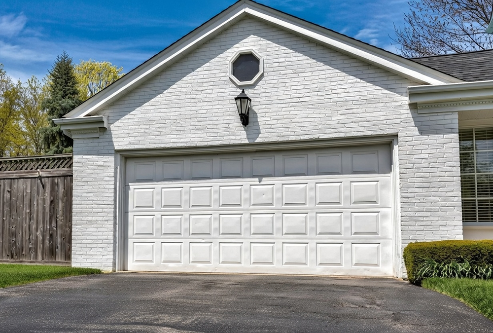 Traditional Garage Door Styles: The Timeless Choice For 2026 Traditional Garage Door Styles Featuring A Clean White Raised-Panel Sectional Overhead Garage Door With Classic Recessed Squares, Mounted On A White Brick Gable-Front Garage, White Trim Frame, Black Lantern Sconce, Pitched Roof, Blue Sky And Spring Greenery In A Suburban Setting, North West Chicago. Lead Contractor Dmytro Shevchenko – Timeless Traditional Garage Door Styles.