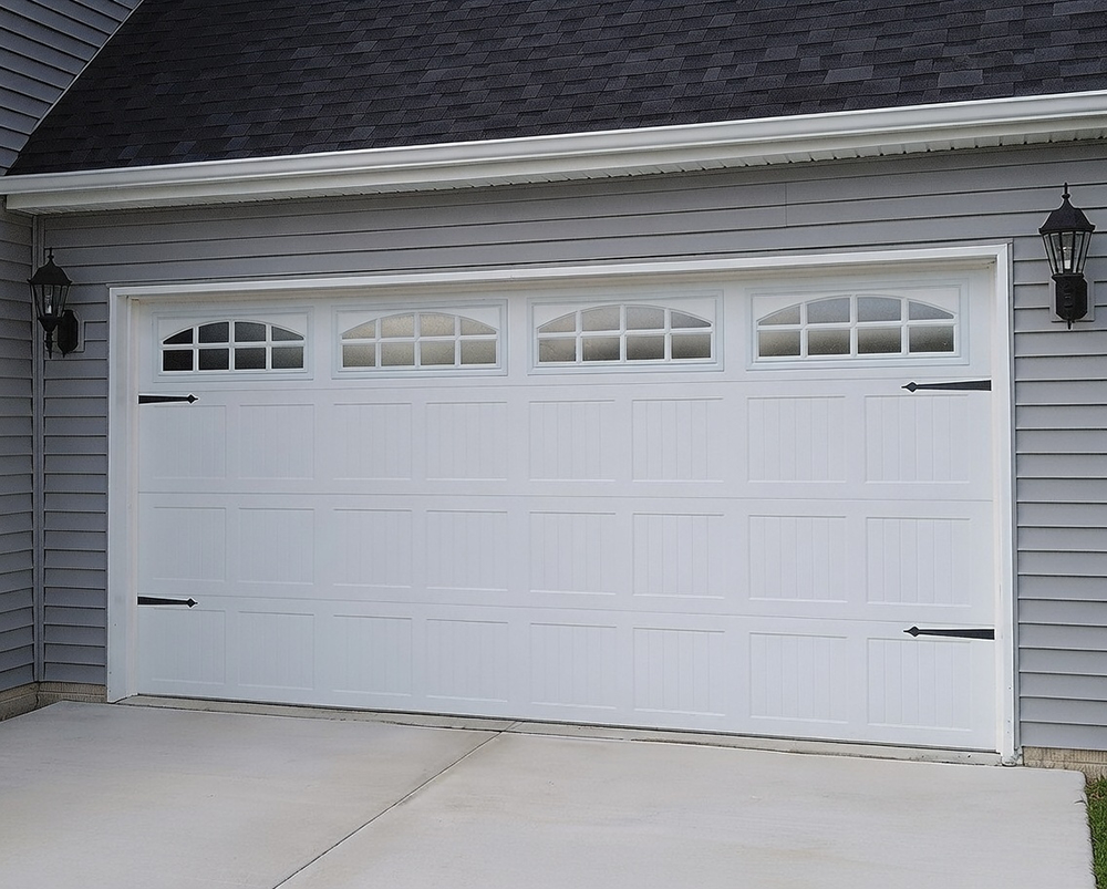 Traditional Garage Door Styles: The Timeless Choice For 2026 Traditional Garage Door Styles Featuring A Pristine White Raised-Panel Sectional Overhead Garage Door With Four Arched Multi-Pane Windows Across The Top Row, Vertical Board-And-Batten Lower Panels, Black Wrought-Iron Decorative Strap Hinges, Clean White Trim Frame, Installed On A Home With Gray Siding And Dark Shingle Roof, Flanked By Black Lantern Sconces, North West Chicago. Lead Contractor Dmytro Shevchenko – Timeless Traditional Garage Door Styles.