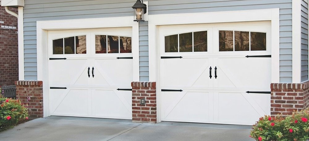 Classic White Carriage-Style Double Garage Doors With Arched Tops, Multiple Grid Glass Windows, Black Wrought-Iron Strap Hinges And Handles, X-Pattern Raised Panels, Installed On A Light Gray Siding Home With Brick Accents In North West Chicago. Garage Door Installation By Lead Contractor Dmytro Shevchenko.