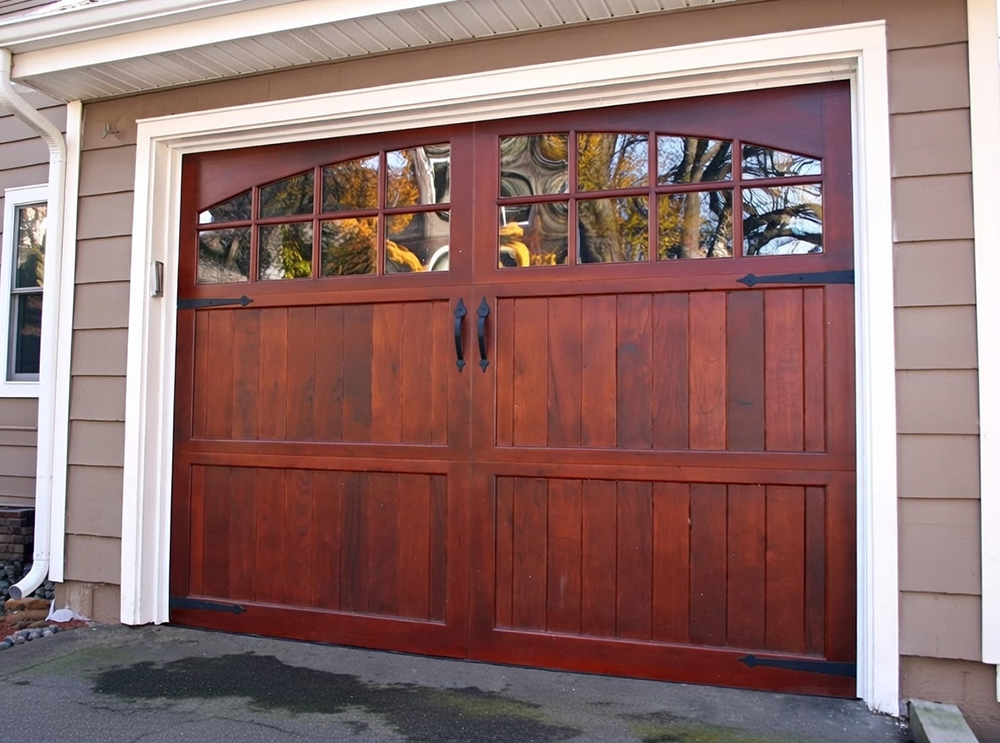 What Are The 5 Types Of Garage Doors? A 2026 Homeowner’s Guide Rich Mahogany Wooden Carriage-Style Swing-Out Garage Doors With Arched Six-Pane Glass Windows, Vertical Board-And-Batten Lower Panels, Black Wrought-Iron Strap Hinges And Decorative Handles, Mounted On A Beige Siding Home With White Trim Frame, Autumn Foliage In Background – Classic Example Of What Are The 5 Types Of Garage Doors. Lead Contractor Dmytro Shevchenko, North West Chicago.