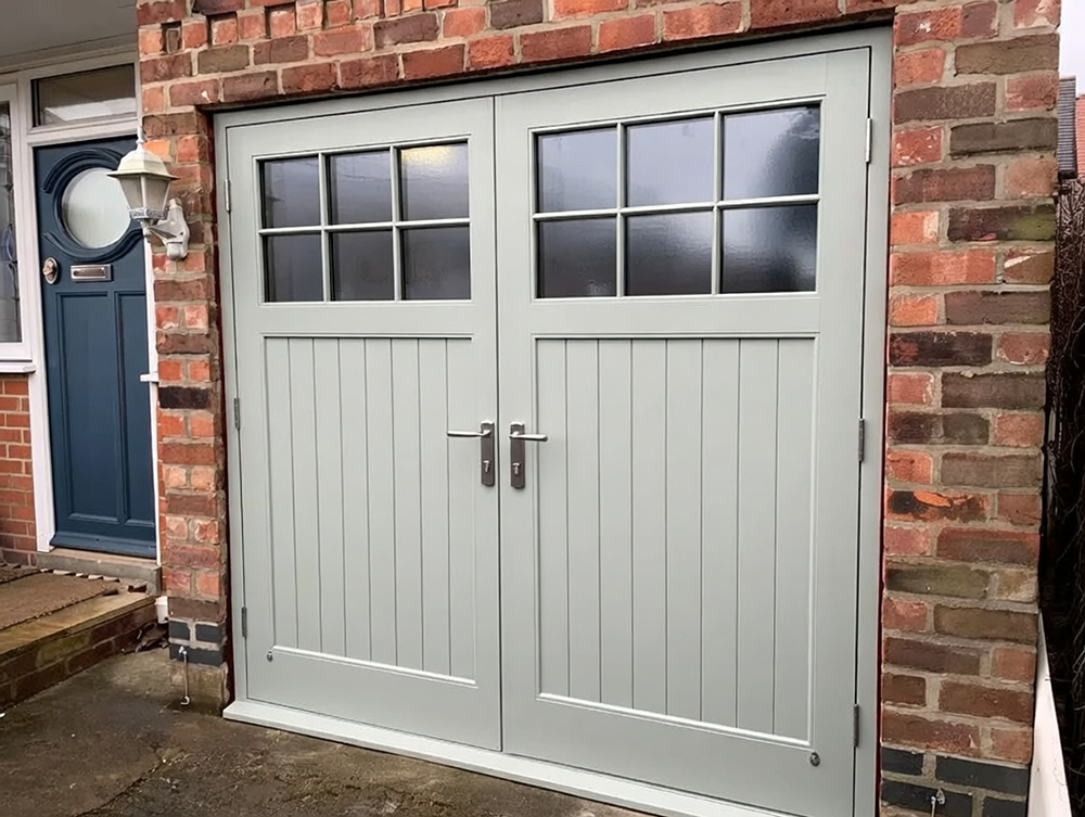 What Are The 5 Types Of Garage Doors? A 2026 Homeowner’s Guide Charming Light Sage Green Carriage-Style Swing-Out Garage Doors With Vertical Board-And-Batten Panels, Six-Pane Frosted Glass Windows In Each Door, Silver Hardware Including Strap Hinges, Pull Handles And Latches, Framed In White Trim On A Classic Red Brick Home With Side Access Path And Blue Front Door Visible – Example Of What Are The 5 Types Of Garage Doors. Lead Contractor Dmytro Shevchenko, North West Chicago.